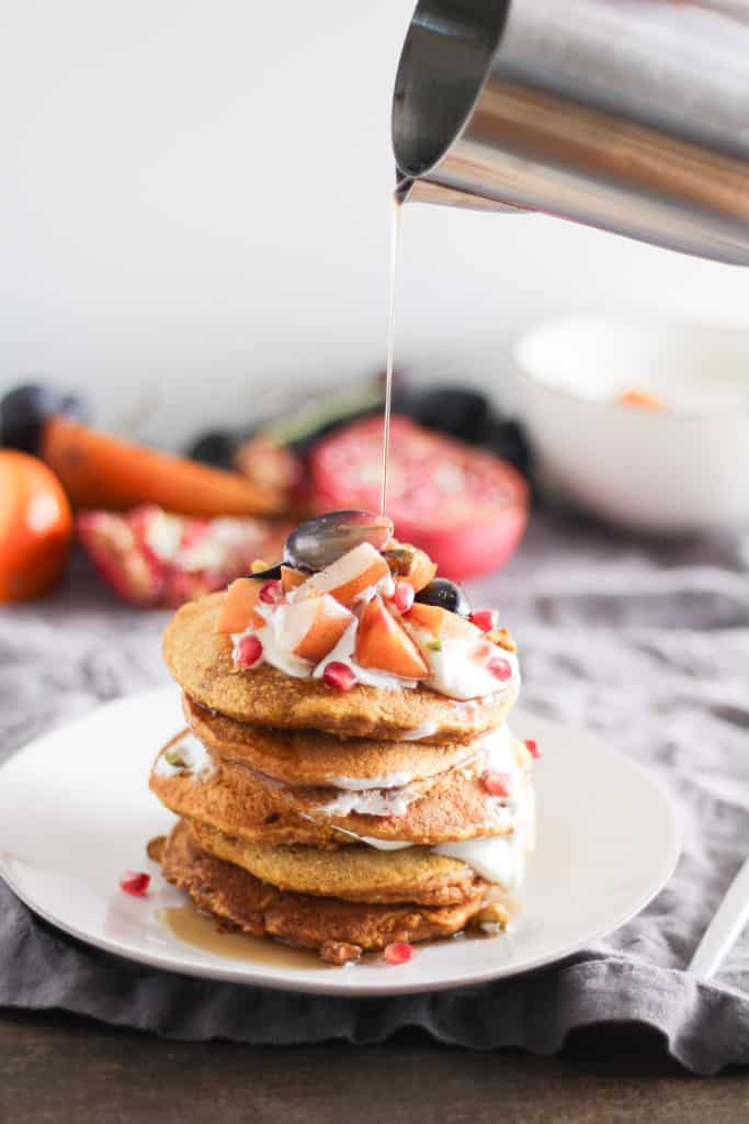 Stack of pumpkin pancakes on white plate with pitcher of syrup pouring over the plate.