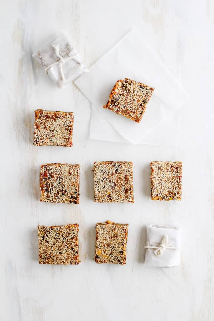Sesame date bars arranged in rows with some wrapped in parchment paper nad tied with string. Set against white backdrop.