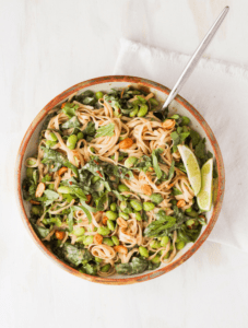 peanut noodles with edamame and spinach in brown dish against white backdrop.