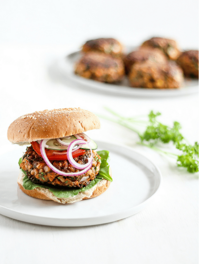 Straight-on shot of Moroccan-spiced chickpea burger on a bun on a white plate with burger patties and herbs in the background.