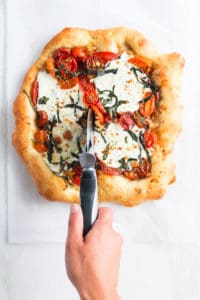 Homemade pizza with caprese toppings and hand with pizza cutter slicing bottom of pizza.