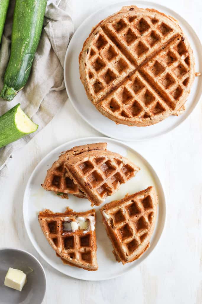 Overhead shot of whole wheat zucchini bread waffles on white plates with zucchini on the side.