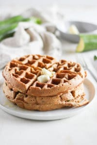 Stack of whole wheat zucchini bread waffles with butter on a white plate with zucchini in background.