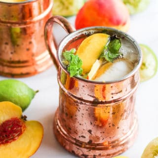Ginger peach kombucha moscow mules in copper mugs on white marble with peaches and lime.