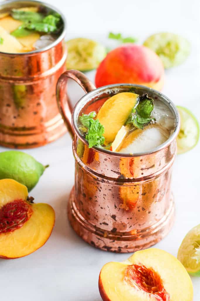 Ginger peach kombucha moscow mules in copper mugs on white marble with peaches and lime.