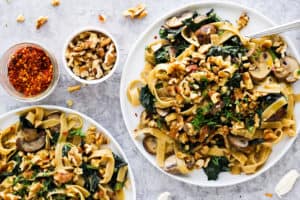 creamy vegan pasta on white plates with small bowls of walnuts and crushed red pepper.