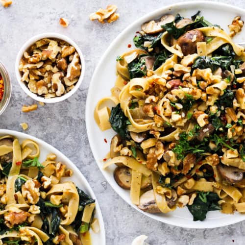 creamy vegan pasta on white plates with small bowls of walnuts and crushed red pepper.