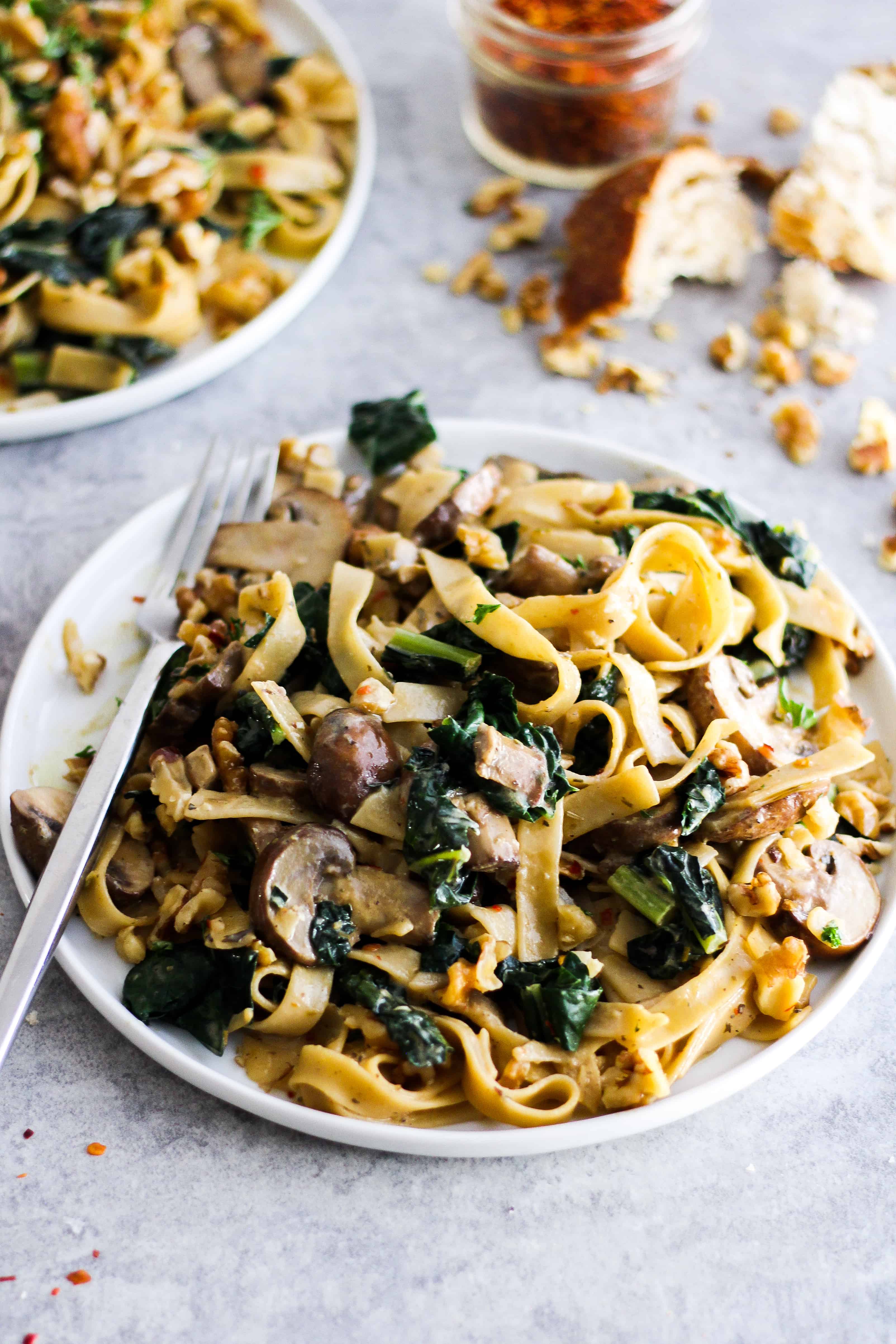 creamy vegan pasta with mushrooms and kale on white plate with bread and crushed red pepper in background.