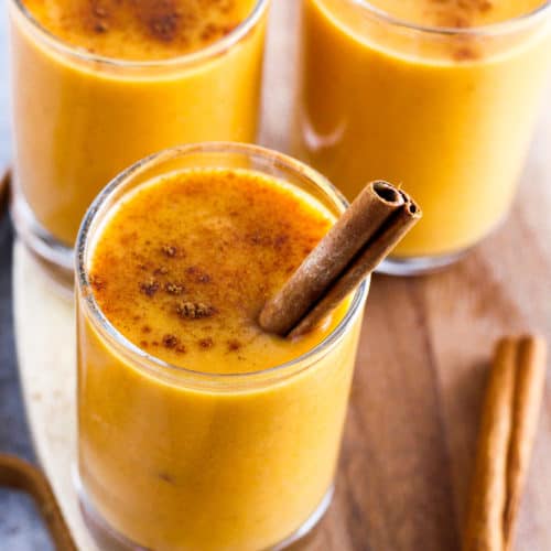 Pumpkin lassi with cinnamon stick in small glass with wood board in background.