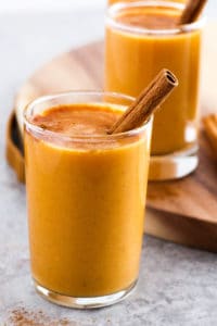 Pumpkin lassi with cinnamon stick in small glass with wood board in background.
