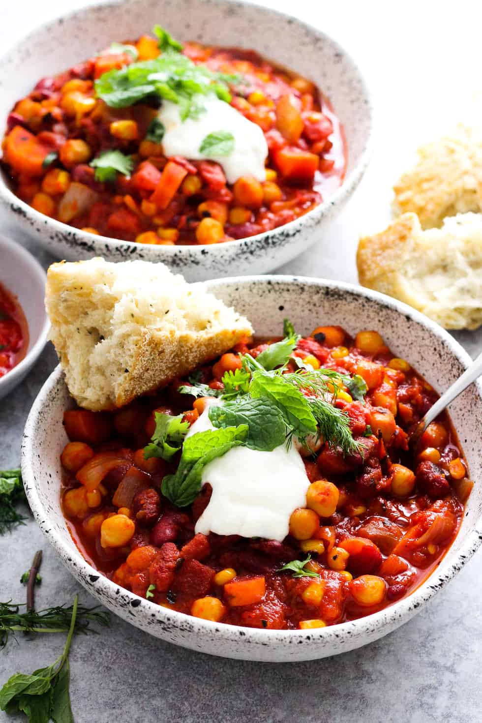 Bean chili with harissa in white stoneware bowls with bread and fresh herbs on top.