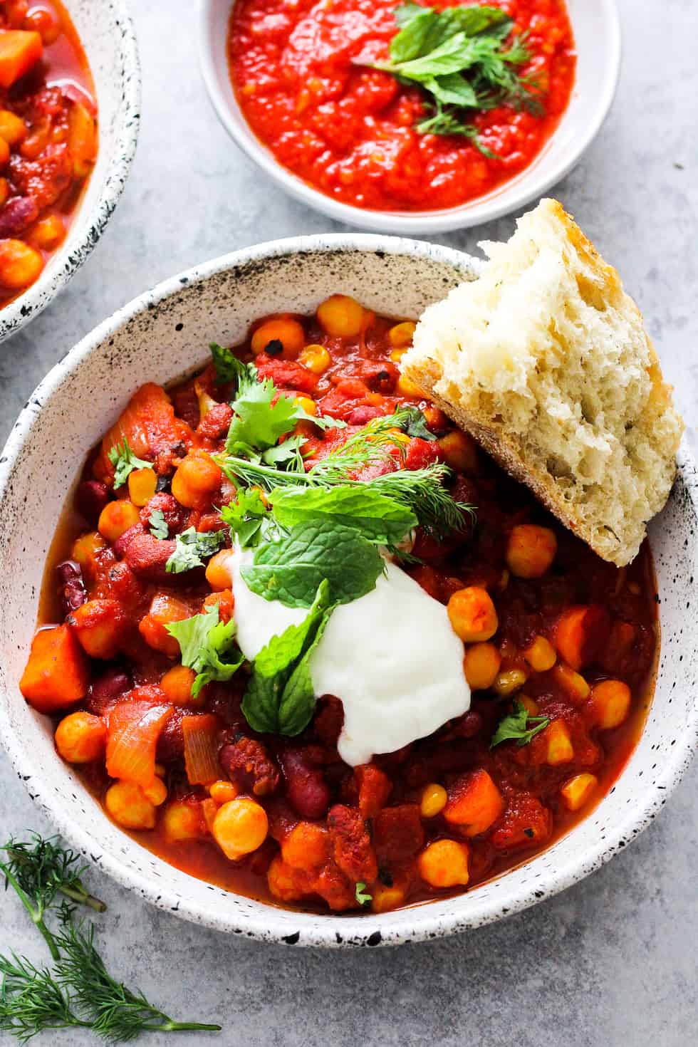 Bean chili with harissa in white stone bowl with yogurt, fresh herbs, and bread on top.