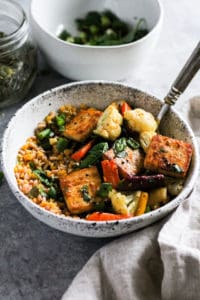 Sheet pan tofu bowls in white stone bowl with herbs and pumpkin seeds in background.