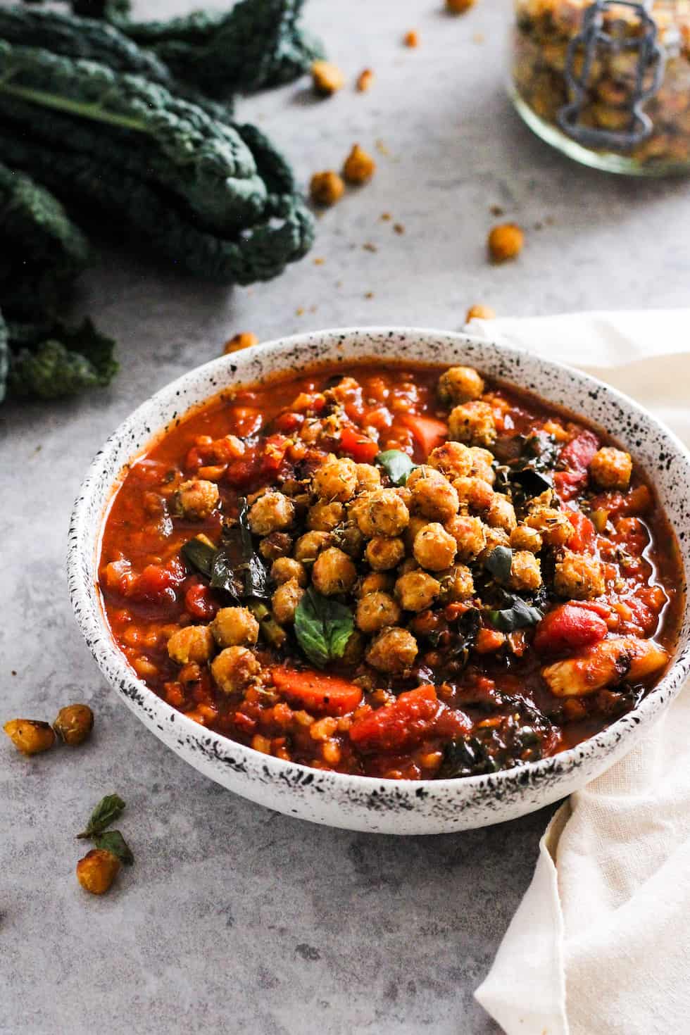 Tomato barley soup with roasted chickpeas in stone bowl with kale and mason jar or chickpeas in the background.