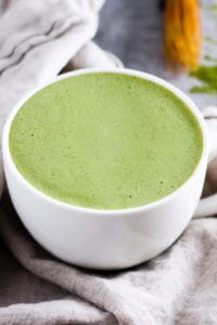 Matcha latte in white mug with cream napkin and whisk in background.