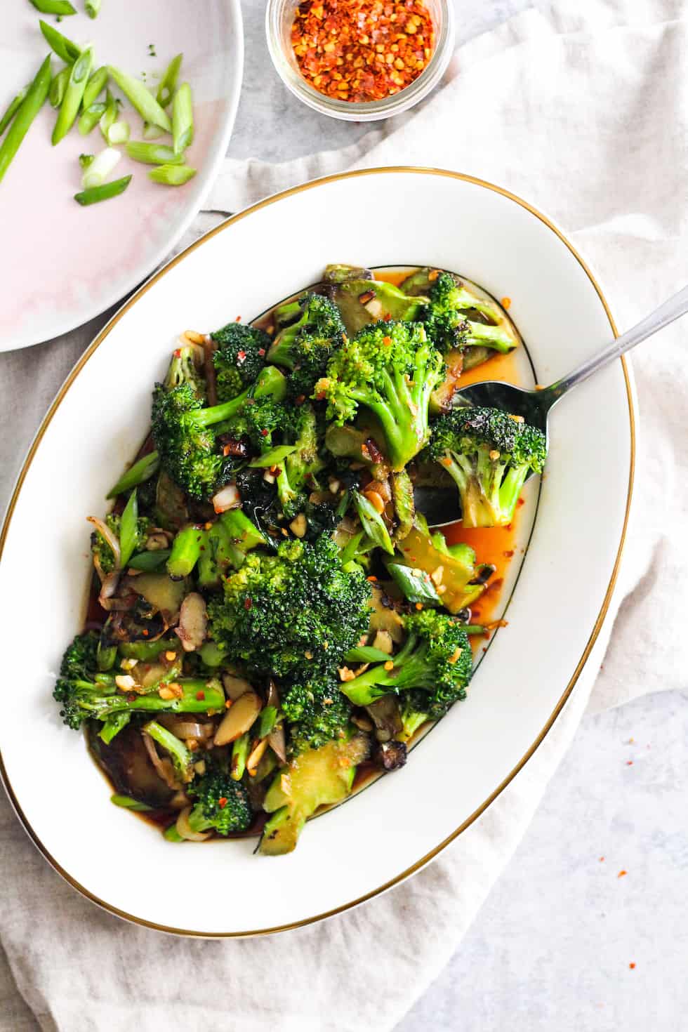 Low waste broccoli stem stir-fry on white platter with plate of green onions and jar of crushed red pepper.