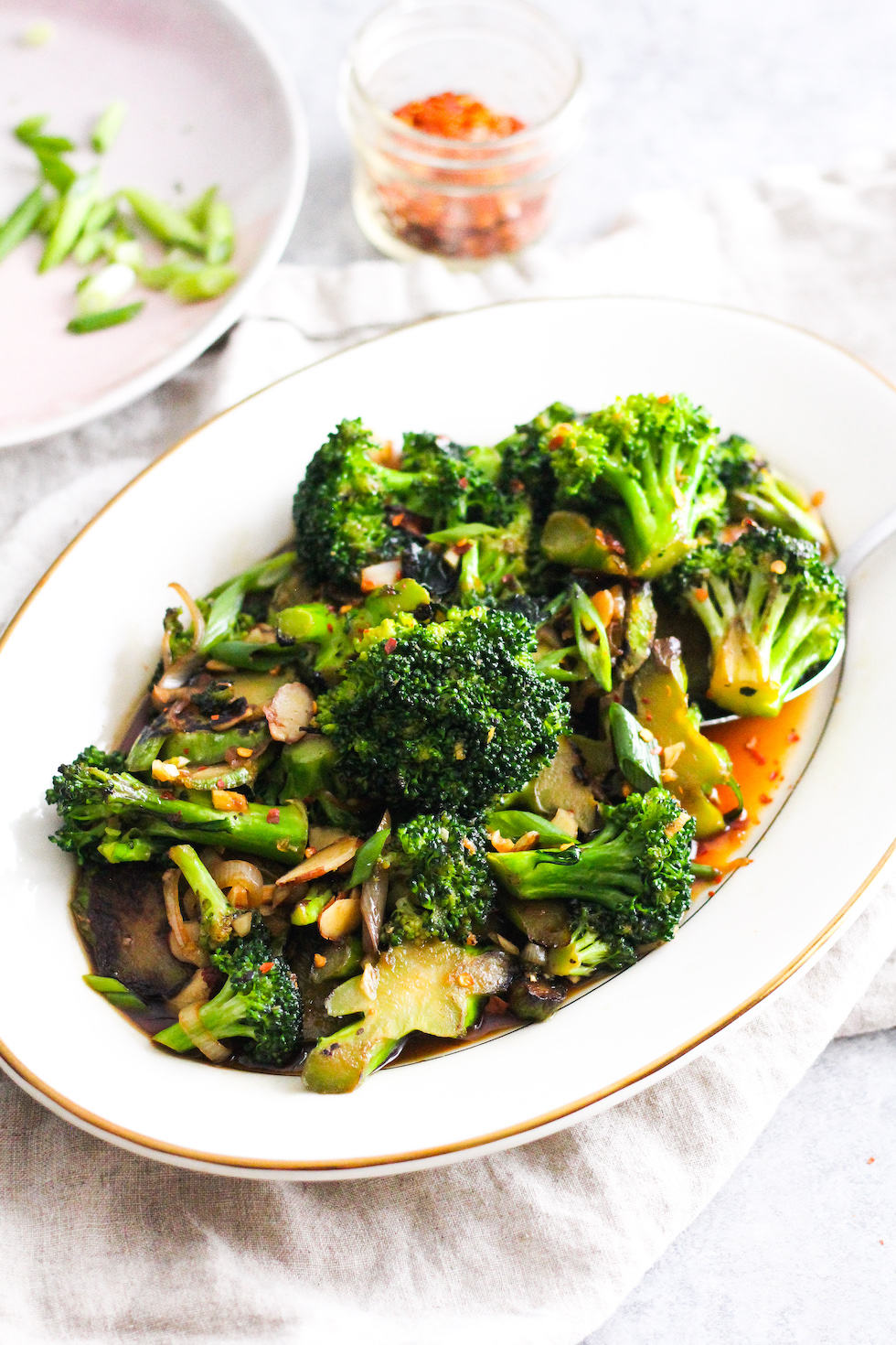 Low waste broccoli stem stir fry on white platter with green onions and crushed red pepper in background.