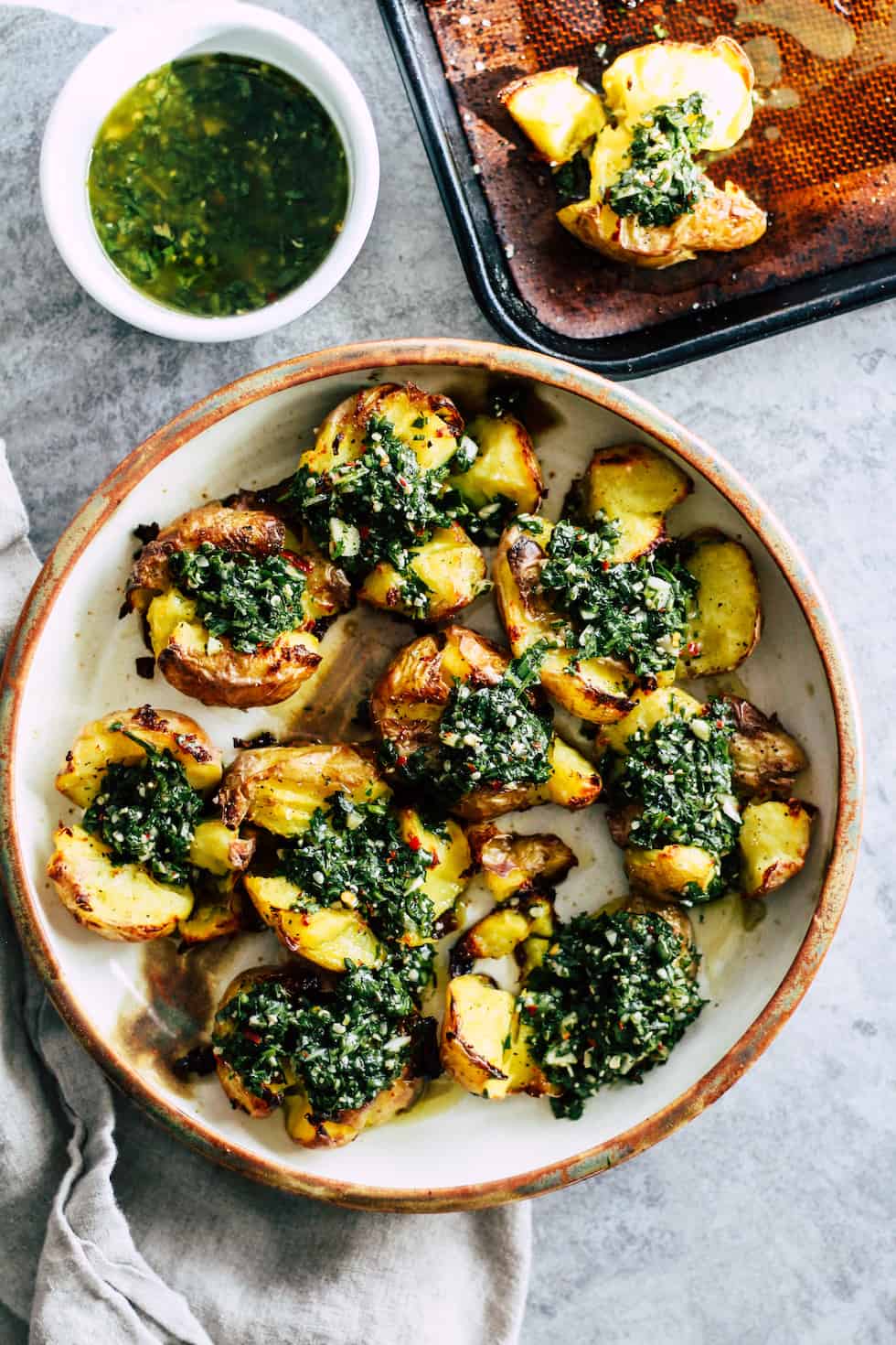 Overhead view of Chimichurri Smashed Potatoes on ceramic dish with small bowl of chimichurri sauce and baking sheet.