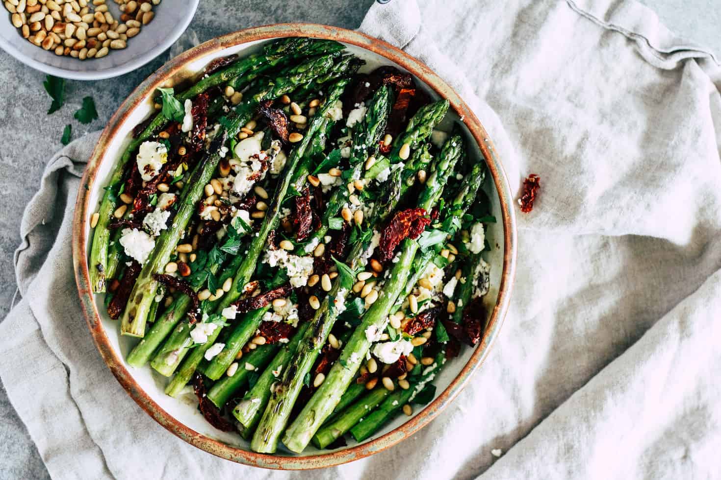 Roasted asparagus in brown ceramic dish with pine nuts and linen napkin.