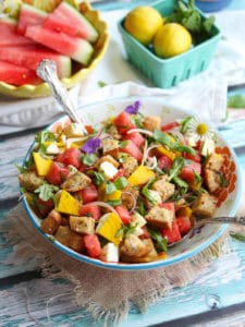 Watermelon panzanella salad in blue bowl with watermelon and carton of lemons in background.