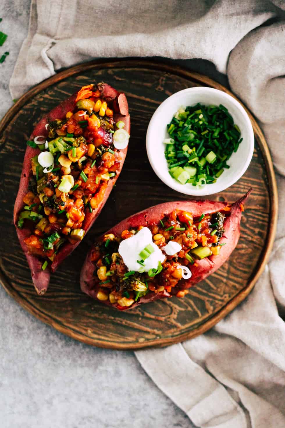 Stuffed sweet potatoes on wood plate with small bowl of chives and green onion.