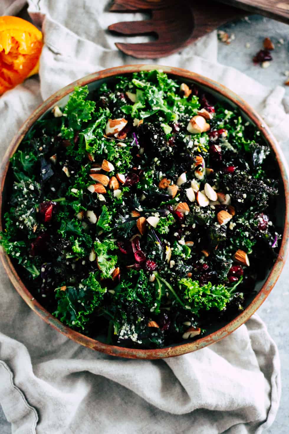 Kale salad in ceramic dish with orange peel and wooden serving spoons.