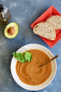 Tomato basil soup in white bowl with avocado and bread.