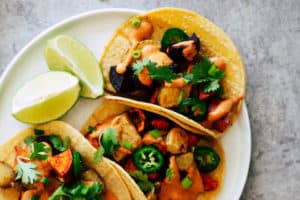 Three vegetable tacos on white plate with lime wedges.