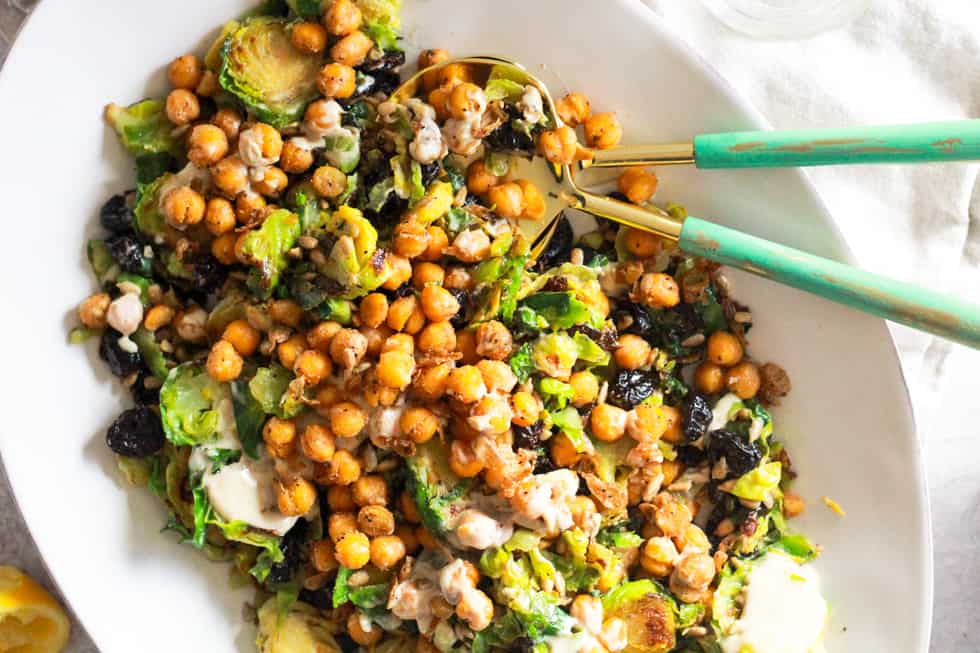 Horizontal image of white platter with Shaved Brussels Sprouts Salad and green serving utensils.