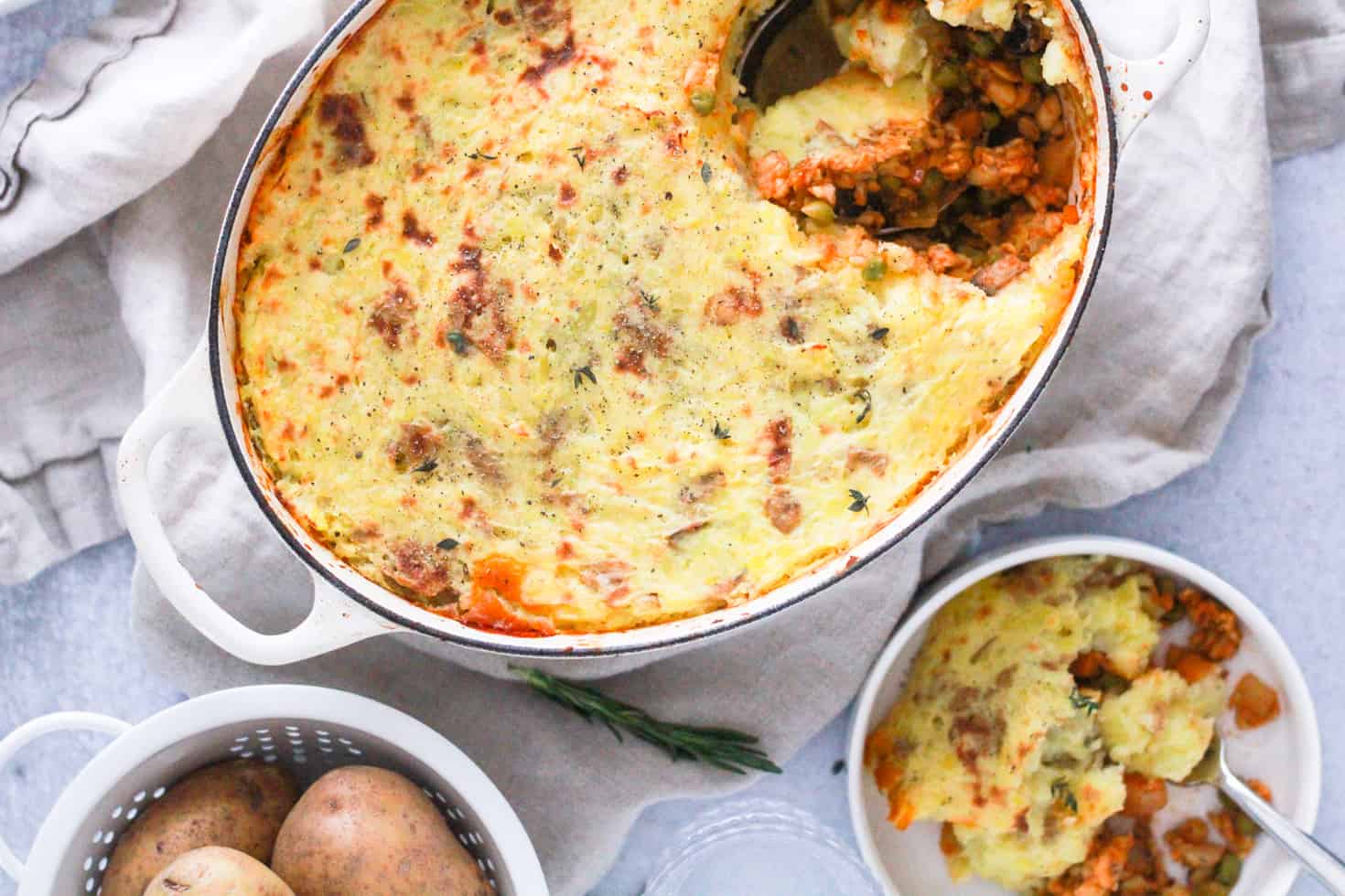 Horizontal image of white dutch oven with tempeh shepherd's pie and gold potatoes in white basket.
