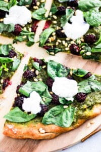 Sliced green Vegan Pesto Pizza with blackberries on wood board.