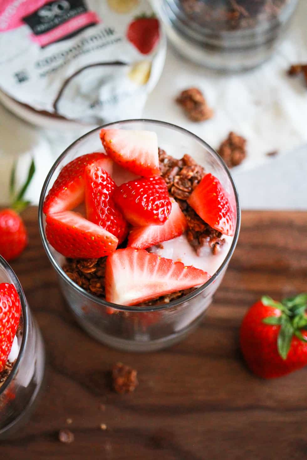 Strawberry yogurt parfait in glass on wood board with carton of So Delicious yogurt.