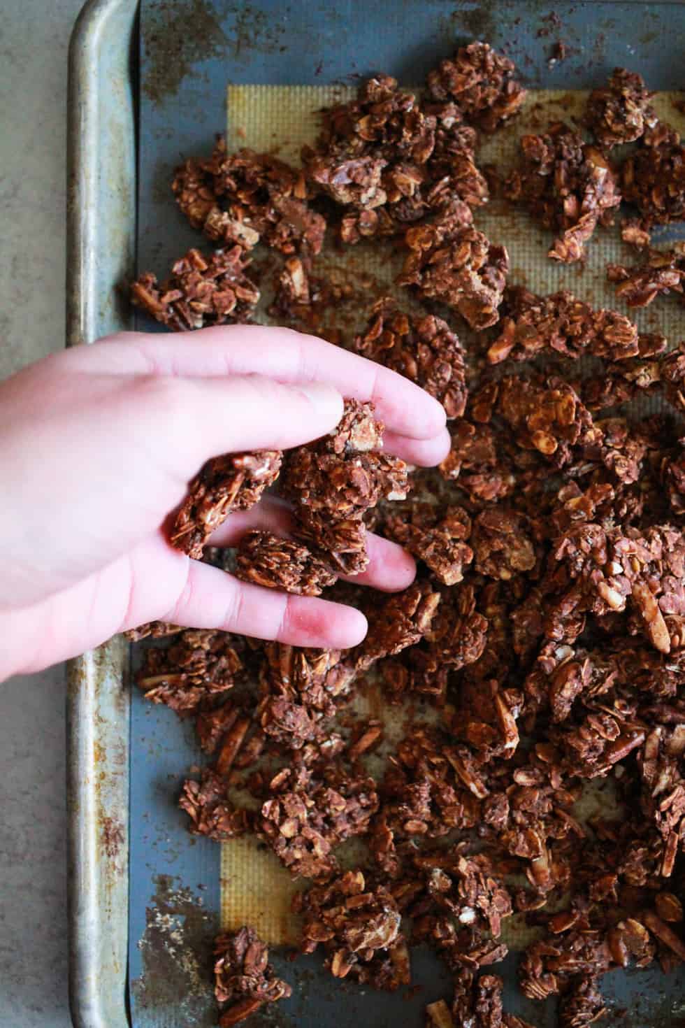 Hand breaking apart chocolate chia seed granola on a baking sheet.