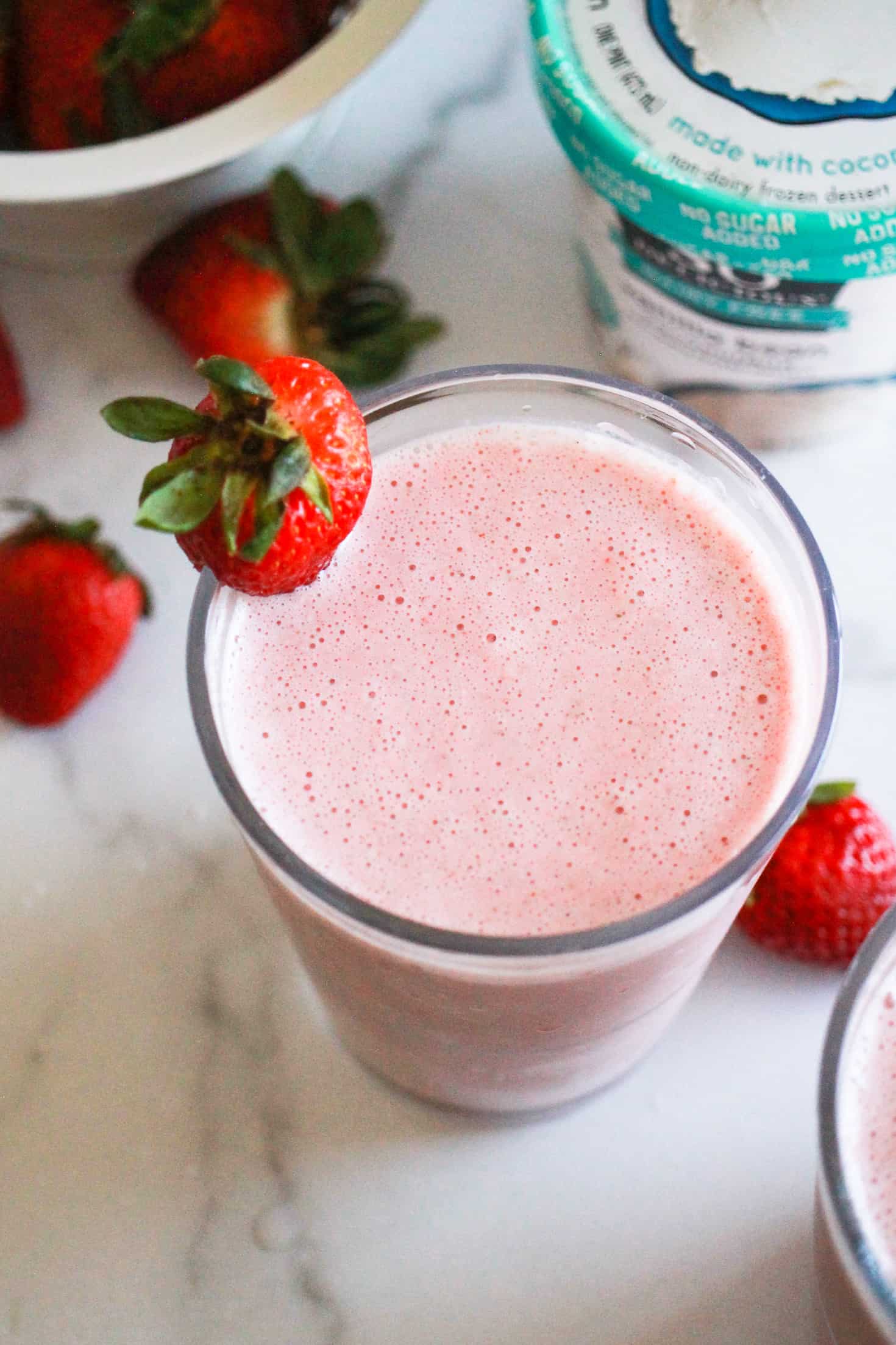 Strawberry milkshake in a glass with So Delicious ice cream alternative and fresh strawberries behind it.