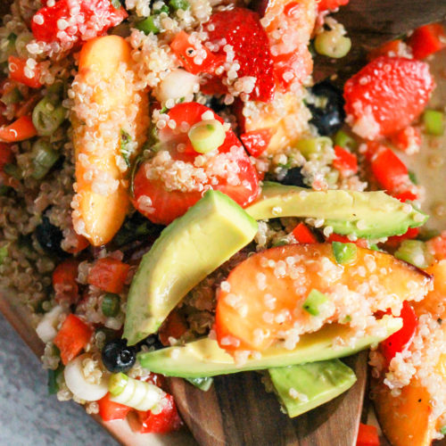 Quinoa salad with avocado and fruit on tan platter with wood spoons.
