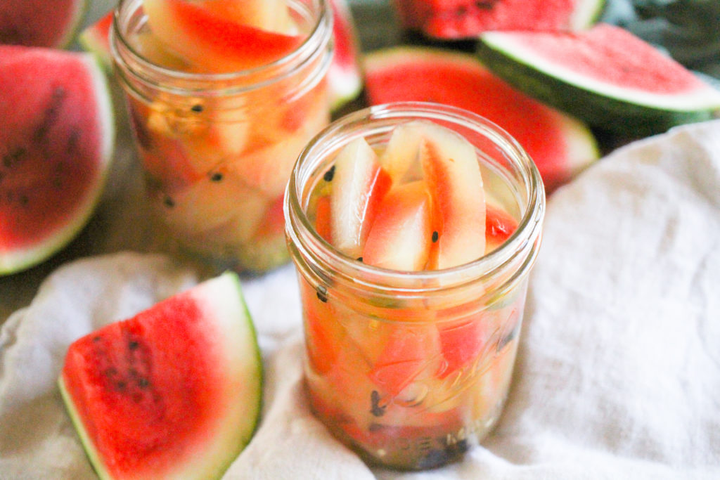 Horizontal image of jars of pickled watermelon rind with watermelon slices around them.