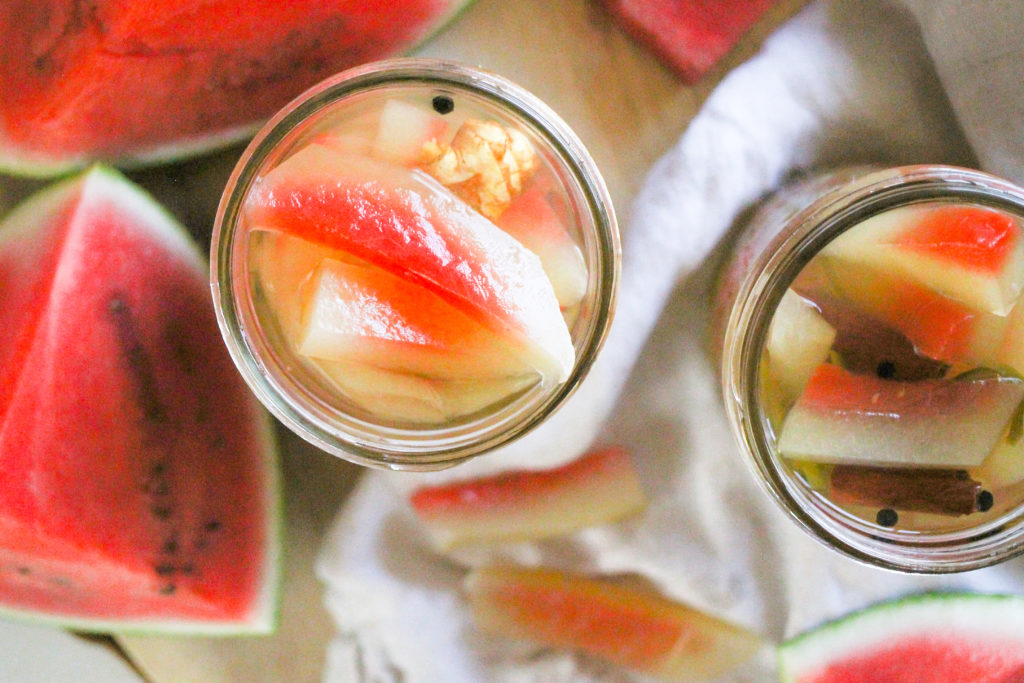 Horizontal overhead image of pickled watermelon rind in glass jars with sliced watermelon around them.