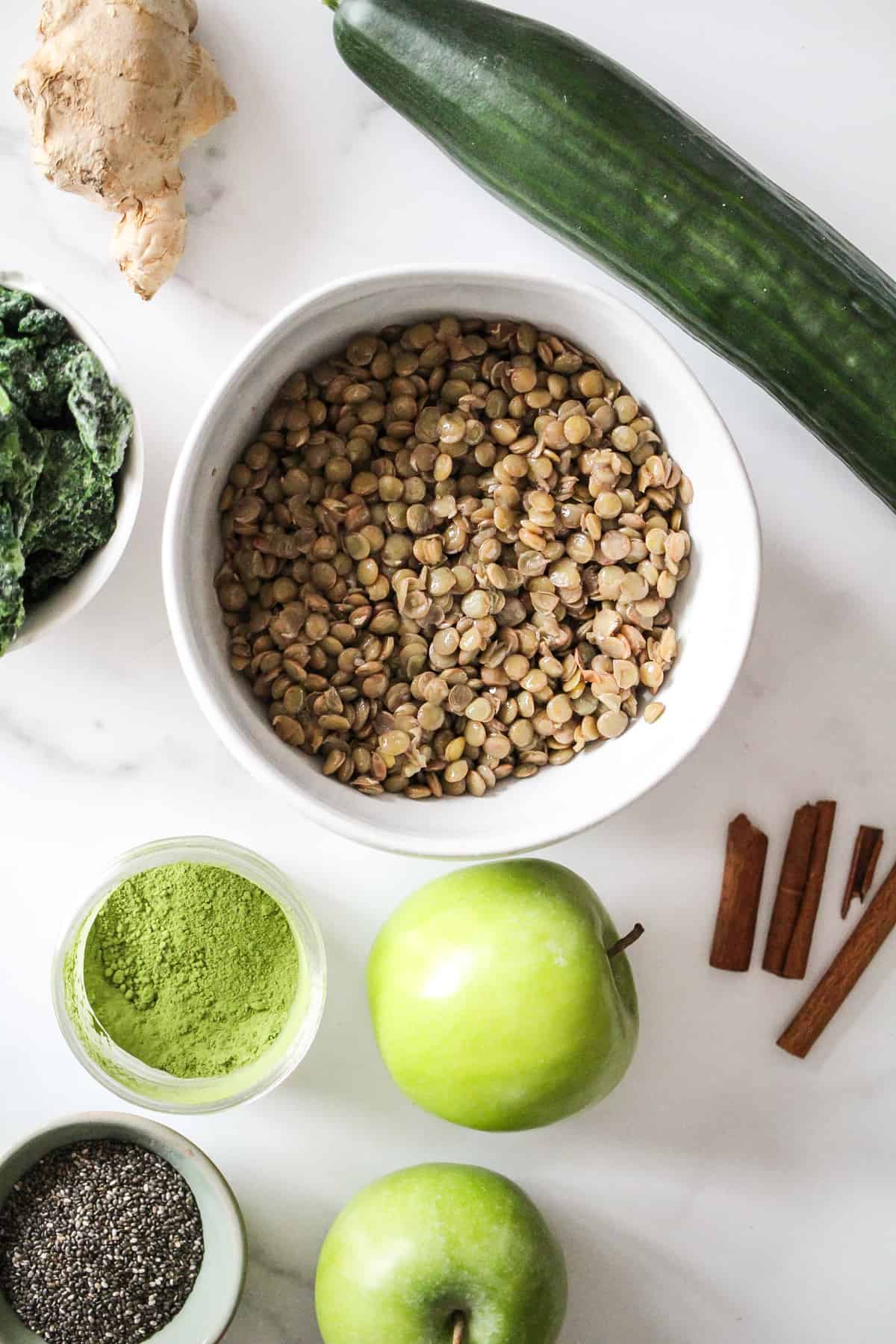 Flatlay image of lentil smoothie ingredients, including cooked lentils, apples, cucumber, cinnamon, ginger, matcha, and chia seeds.