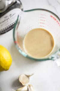 Tahini in a glass measuring cup with garlic, lemon, and maple syrup.