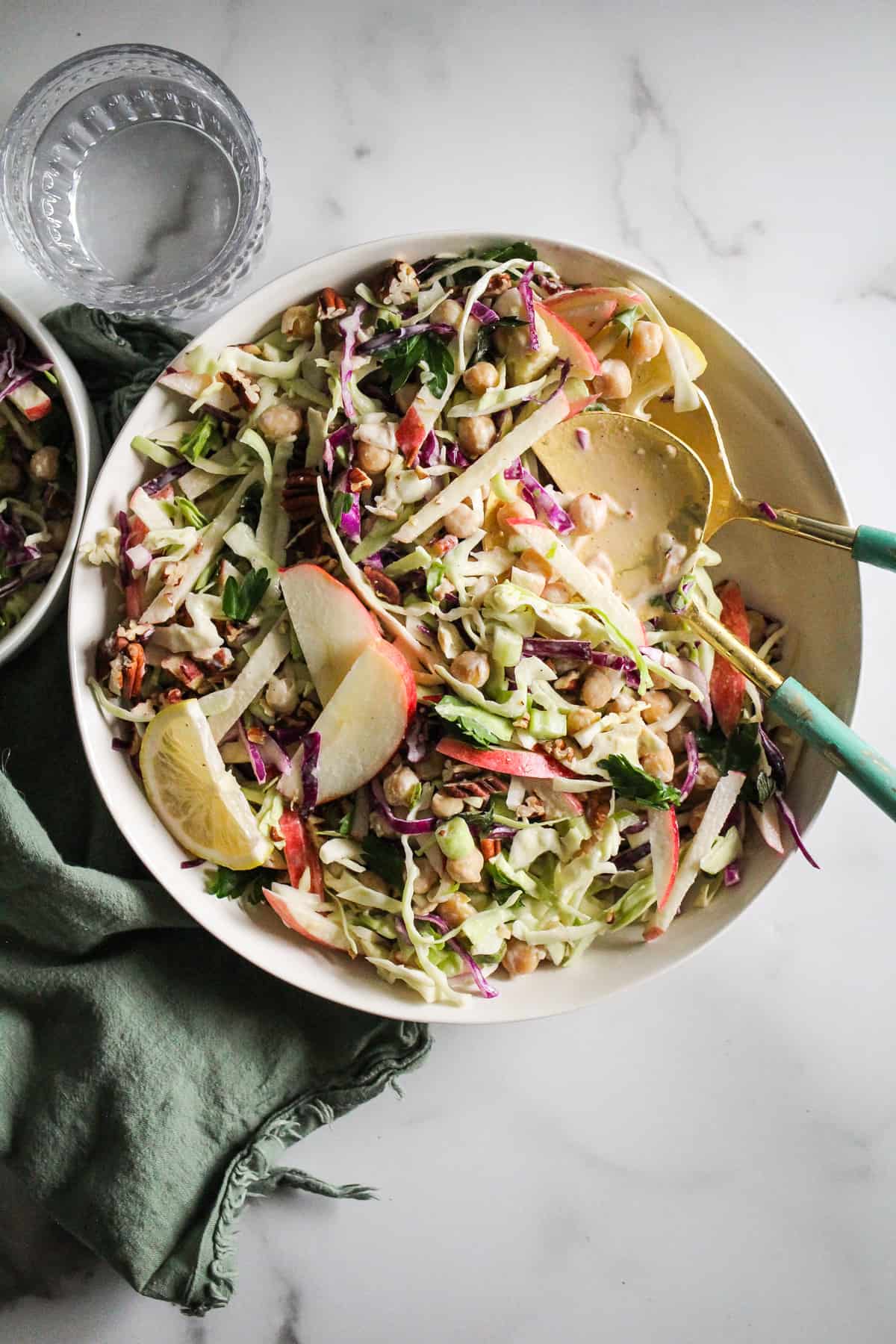Overhead image of vegan coleslaw with apples and lemon tahini in white bowl with blue serving spoons.