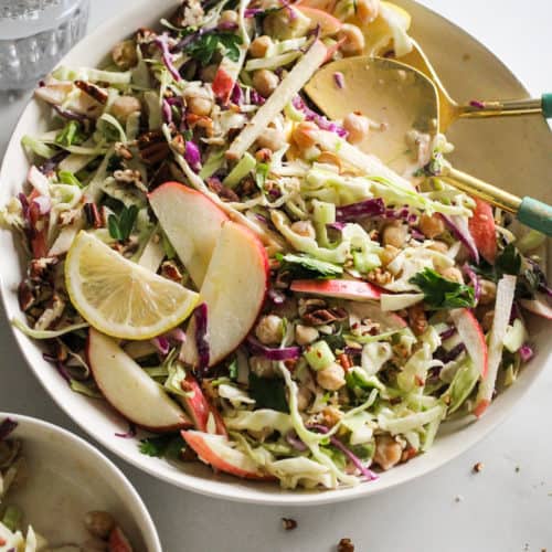 Vegan coleslaw in a white bowl with a glass of water behind it and blue and gold serving spoons.
