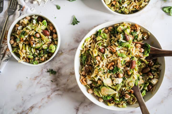 Horizontal image of brussels sprouts lemon orzo salad in white bowls on marble counter.