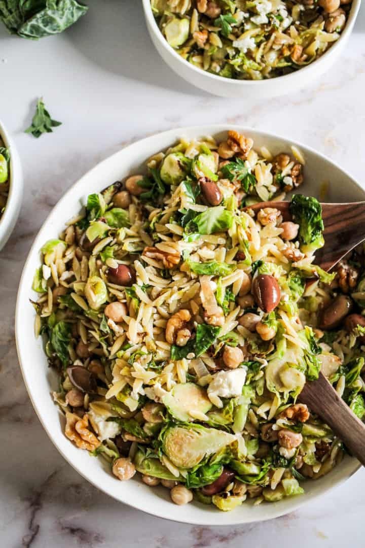 Closeup image of orzo salad with brussels sprouts and olives in a white bowl with wood serving spoons.