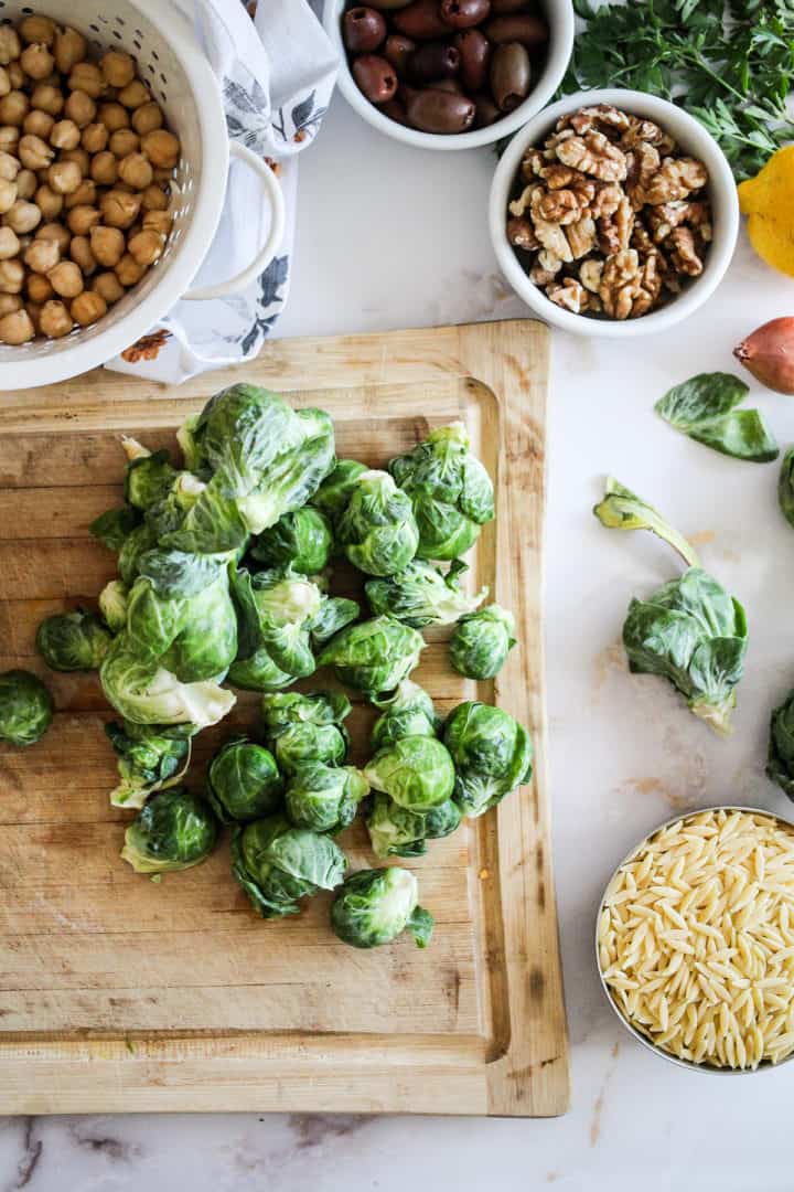 Brussels sprouts on a wood cutting board surrounded by chickpeas, olives, walnuts, and dry orzo pasta.