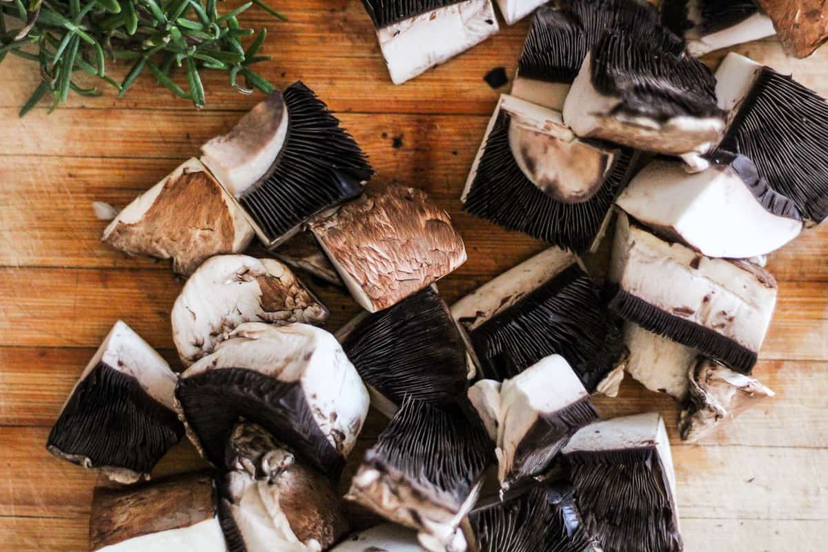 Horizontal image of chopped portobello mushrooms and fresh rosemary sprigs on a wood cutting board.
