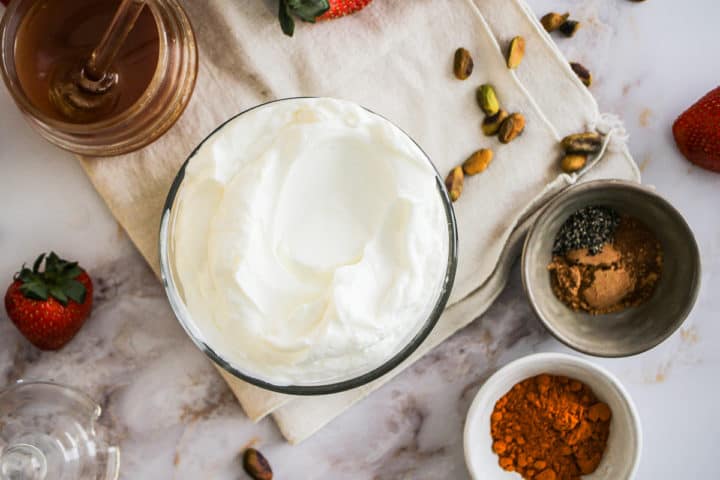 Flatlay image of yogurt bowl ingredients: yogurt, spices, pistachios, honey, and strawberries.