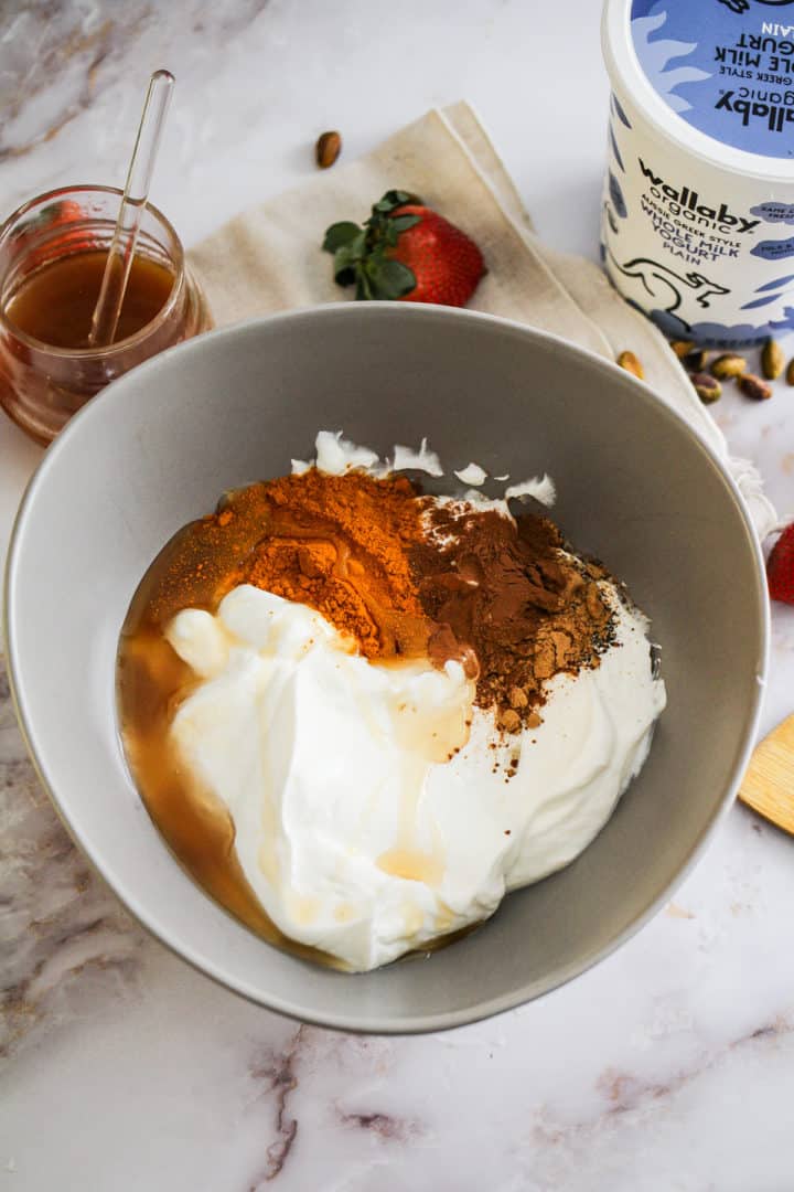 Yogurt, honey, and ground spices in a large grey mixing bowl with a container of yogurt and jar of honey.