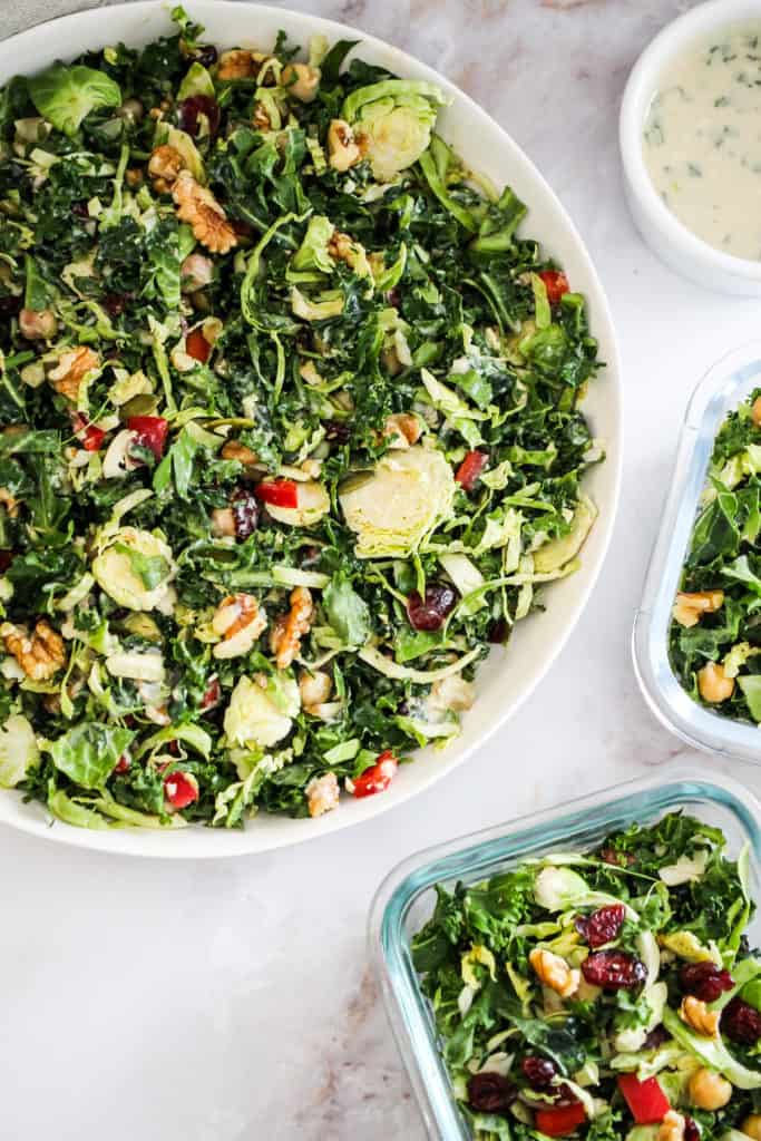 Large white bowl of green salad with two glass meal prep containers filled with salad and a ramekin of tahini dressing.