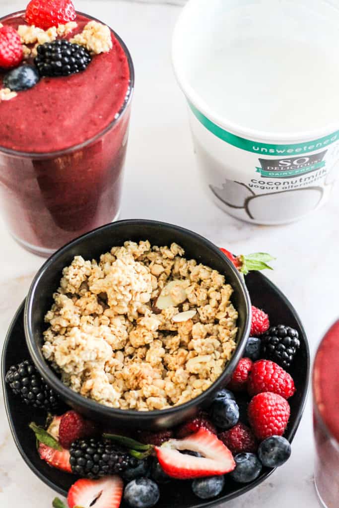 Bowl of granola on a plate of fresh berries with a smoothie and tub of So Delicious yogurt alternative behind it.