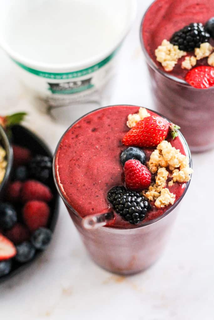 Closeup image of a berry smoothie in a glass with a glass straw garnished with fresh berries and granola with a tub of yogurt alternative behind it.